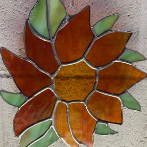 May include: A stained glass sunflower with orange and brown petals and green leaves. The sunflower is made of individual pieces of glass that are joined together with lead.