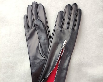 May include: A pair of black leather gloves featuring a silver-toned metal zipper running along the side of the wrist. The interior lining of the gloves is a vibrant red fabric, which is visible where the zipper is partially opened.