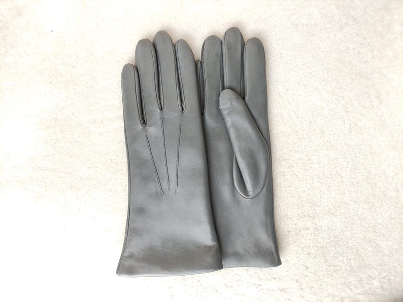 Peut inclure: Une paire de gants en cuir gris mat et lisse. Le gant gauche montre le dos avec trois lignes de couture verticales partant des doigts vers le poignet, tandis que le gant droit pr&eacute;sente le c&ocirc;t&eacute; paume avec une section pour le pouce.
