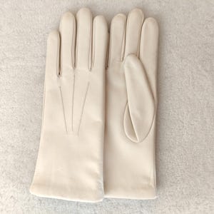 Peut inclure: Une paire de gants en cuir de couleur cr&egrave;me avec une finition lisse et trois lignes de couture d&eacute;coratives verticales sur le dos de la main. Les gants pr&eacute;sentent une coupe classique avec une ouverture droite au poignet et des doigts s&eacute;par&eacute;s.
