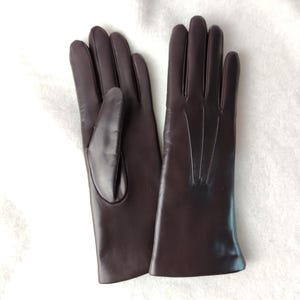 Peut inclure: Une paire de gants marron fonc&eacute; &agrave; la finition lisse. Un gant montre le c&ocirc;t&eacute; paume avec un gousset pour le pouce, tandis que l'autre affiche le dos avec trois lignes verticales cousues partant de la base des doigts vers le bas.