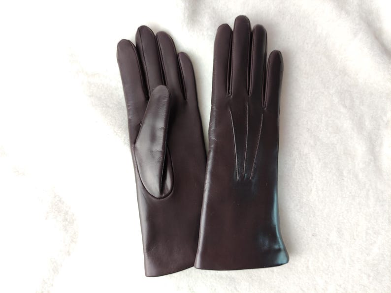 Peut inclure: Une paire de gants marron fonc&eacute; &agrave; la finition lisse. Un gant montre le c&ocirc;t&eacute; paume avec un gousset pour le pouce, tandis que l'autre affiche le dos avec trois lignes verticales cousues partant de la base des doigts vers le bas.