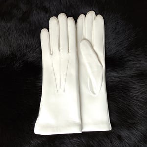 Peut inclure: Une paire de gants en cuir blanc avec quatre doigts et un pouce, pr&eacute;sentant trois lignes de couture verticales sur le dos de la main. Les gants ont une finition lisse et d&eacute;passent le poignet, affichant une couleur blanche unie et nette.