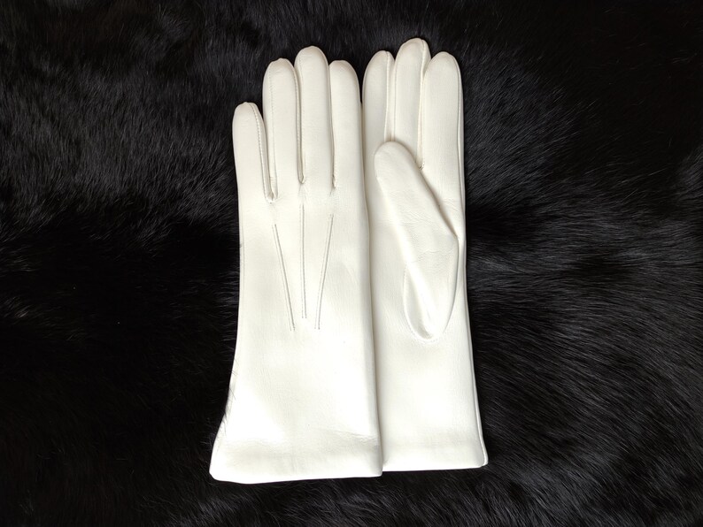 Peut inclure: Une paire de gants en cuir blanc avec quatre doigts et un pouce, pr&eacute;sentant trois lignes de couture verticales sur le dos de la main. Les gants ont une finition lisse et d&eacute;passent le poignet, affichant une couleur blanche unie et nette.
