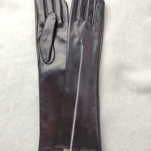 May include: A pair of long black leather gloves featuring a vertical silver-toned metal zip running down the centre of the forearm. The gloves have a smooth, polished finish and extend past the wrist, showcasing a sleek, fitted design.