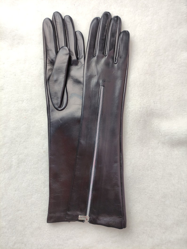 May include: A pair of long black leather gloves featuring a vertical silver-toned metal zip running down the centre of the forearm. The gloves have a smooth, polished finish and extend past the wrist, showcasing a sleek, fitted design.