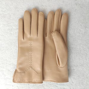 Puede incluir: Un par de guantes de cuero color beige claro, colocados sobre una superficie blanca. Los guantes tienen un dise&ntilde;o cl&aacute;sico con detalles cosidos a lo largo de los dedos y la mu&ntilde;eca. El cuero parece liso y flexible.