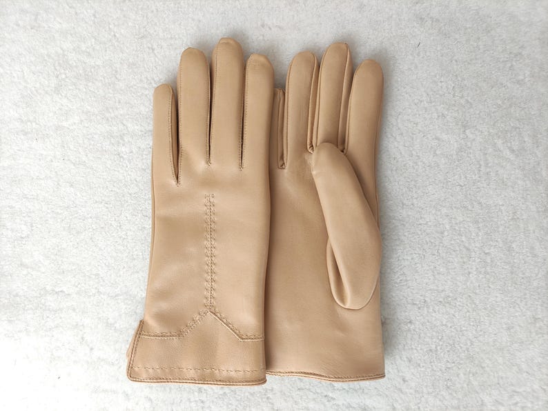 Puede incluir: Un par de guantes de cuero color beige claro, colocados sobre una superficie blanca. Los guantes tienen un dise&ntilde;o cl&aacute;sico con detalles cosidos a lo largo de los dedos y la mu&ntilde;eca. El cuero parece liso y flexible.