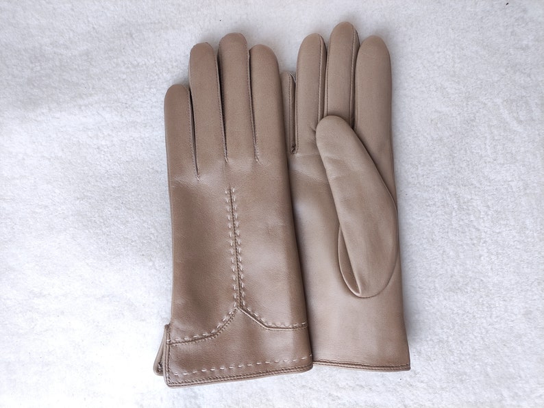 Puede incluir: Un par de guantes de cuero color canela. Los guantes est&aacute;n hechos de cuero liso y presentan costuras decorativas a lo largo de las costuras y en la mu&ntilde;eca. Los guantes est&aacute;n colocados sobre una superficie blanca.