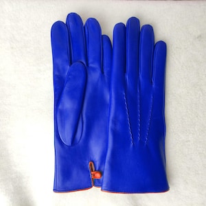 May include: A pair of vibrant royal blue gloves featuring a smooth finish, four fingers, and a thumb. The wrist area includes a small orange button closure detail with matching orange piping along the bottom edge of the cuffs.
