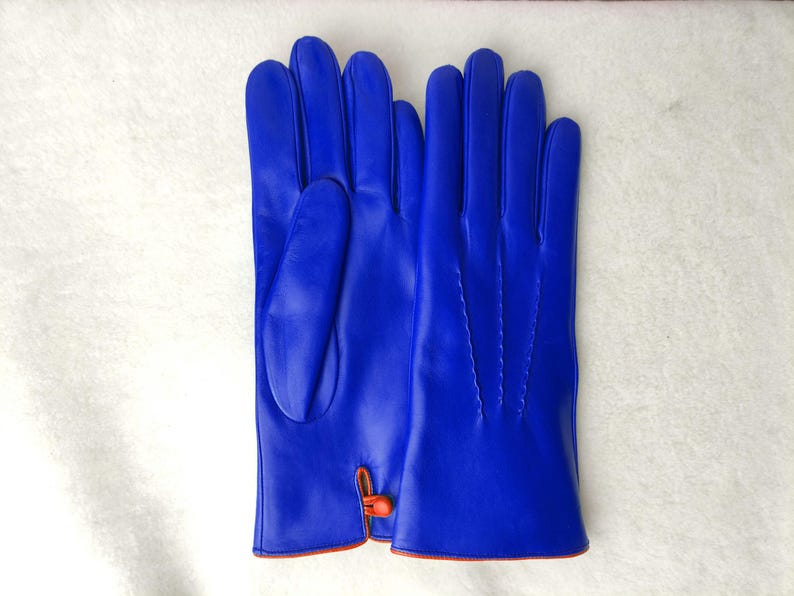 May include: A pair of vibrant royal blue gloves featuring a smooth finish, four fingers, and a thumb. The wrist area includes a small orange button closure detail with matching orange piping along the bottom edge of the cuffs.