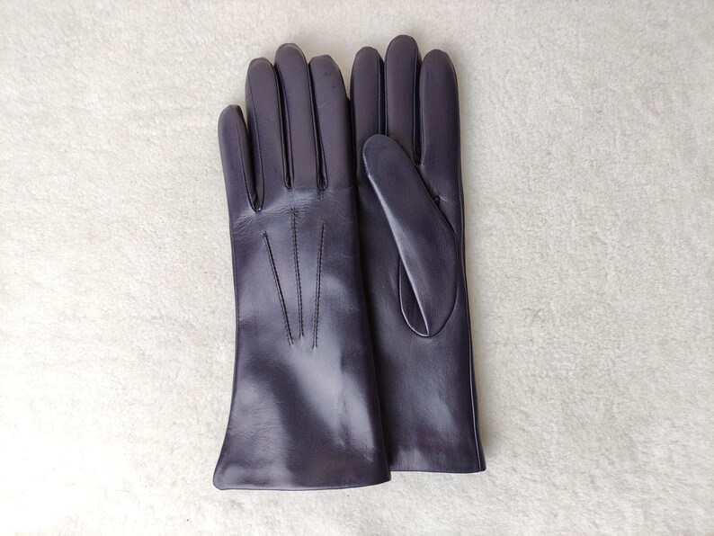 Peut inclure: Une paire de gants en cuir violet fonc&eacute; avec une finition lisse, quatre doigts et un pouce. Le dos du gant gauche pr&eacute;sente trois lignes de couture verticales s'&eacute;tendant de la base des doigts vers la zone du poignet.