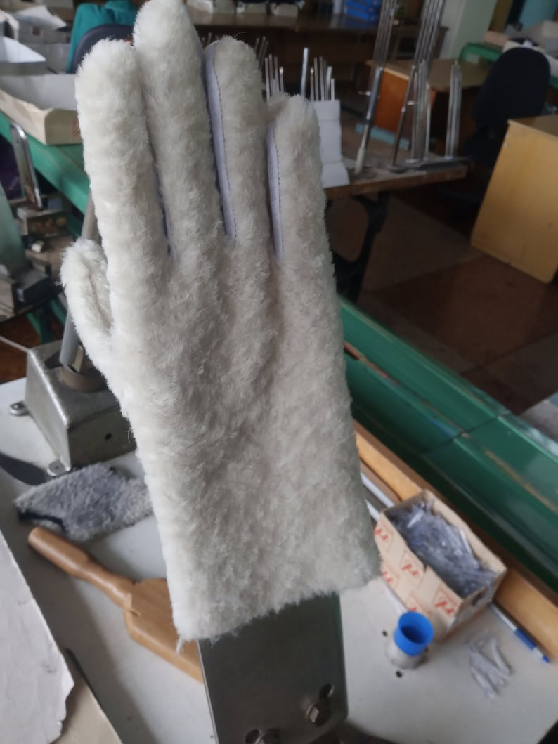 May include: A white, fluffy glove with a leather palm and white stitching. The glove is made of a soft, textured material and appears to be new. The glove is designed to fit a right hand.