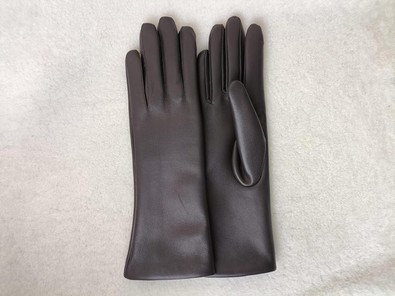 May include: A pair of dark brown gloves with a smooth, matte finish. The gloves feature a classic design with four fingers and a thumb section, extending to the wrist. The material appears uniform in color and texture throughout the surface.