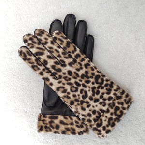 以下が含まれることがあります： 黒い革手袋。手の甲と袖口にヒョウ柄のデザイン。ヒョウ柄は、ベージュの背景に茶色と黒の斑点があります。手袋は白い表面に置かれています。
