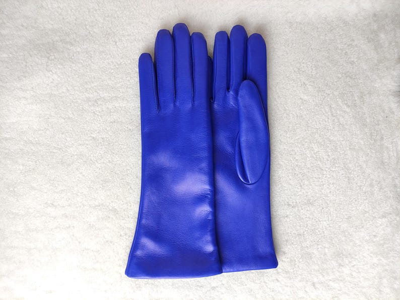 May include: A pair of vibrant royal blue leather gloves featuring a smooth finish, long cuffs, and individual finger compartments. The gloves are displayed side by side, showcasing the consistent saturated blue color and the clean, structured seams of the material.
