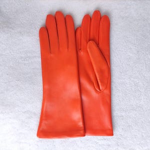 May include: A pair of vibrant orange leather gloves featuring a smooth finish, four fingers, and a thumb on each hand. The gloves have a classic cut with a straight wrist opening, showcasing a solid, bright orange color throughout the design.
