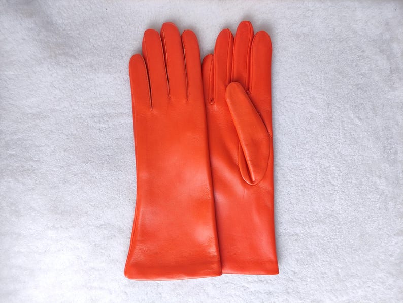 May include: A pair of vibrant orange leather gloves featuring a smooth finish, four fingers, and a thumb on each hand. The gloves have a classic cut with a straight wrist opening, showcasing a solid, bright orange color throughout the design.