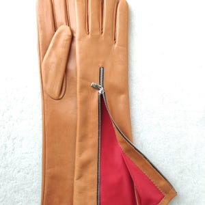 May include: A pair of tan leather gloves featuring a silver-toned metal zip on the side of the wrist. The interior lining is a vibrant red fabric, and a small white tag inside the cuff displays the size 8.5 in black text.
