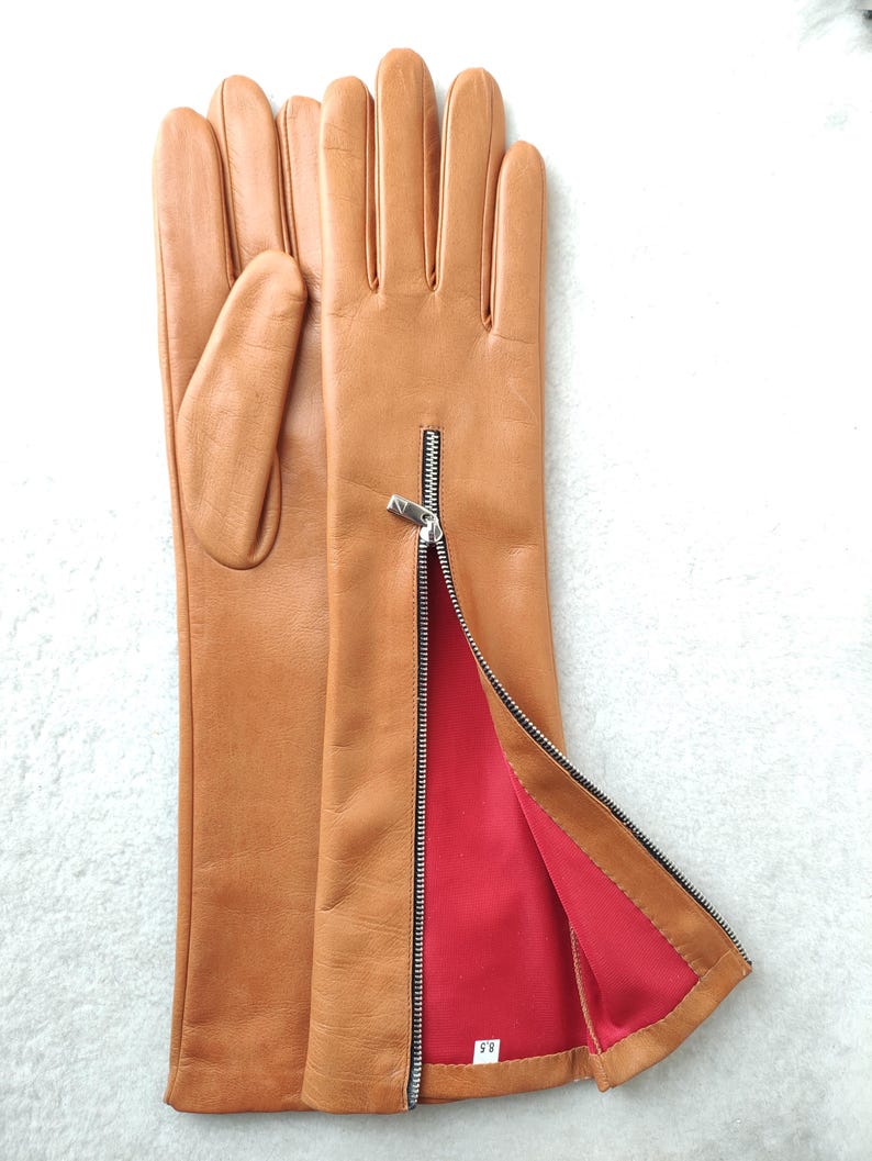 May include: A pair of tan leather gloves featuring a silver-toned metal zip on the side of the wrist. The interior lining is a vibrant red fabric, and a small white tag inside the cuff displays the size 8.5 in black text.