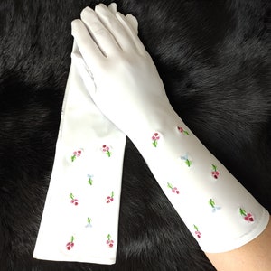 May include: A pair of white long gloves with embroidered pink and blue flowers. The gloves are on a black fur surface.