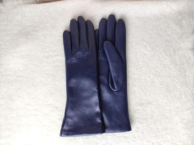 May include: A pair of dark purple leather gloves with a smooth finish. The gloves feature a classic design with four fingers and a thumb, extending past the wrist. The leather has a subtle sheen and visible natural grain texture throughout.