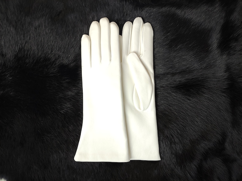May include: A pair of solid white gloves with a smooth, matte finish. The gloves feature a classic design with four fingers and a thumb, extending past the wrist with a slightly flared opening at the cuff, showcasing clean, stitched seams.