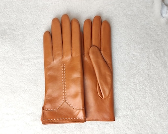May include: A pair of tan leather gloves featuring decorative stitching along the back of the hand and a horizontal stitched detail across the wrist area. The gloves have a smooth finish and are displayed side by side on a textured surface.
