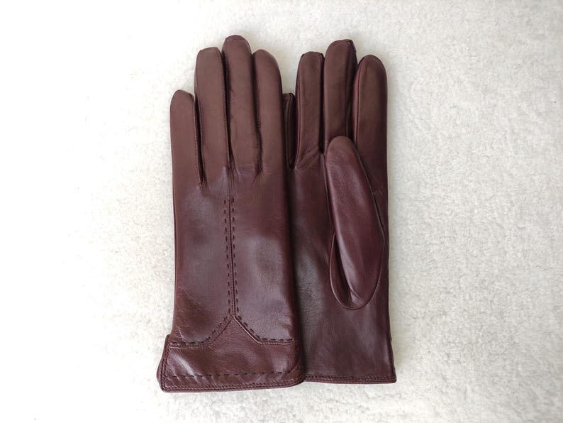 Puede incluir: Un par de guantes de cuero burdeos. Los guantes est&aacute;n hechos de cuero liso y presentan costuras decorativas a lo largo de las costuras. Los guantes est&aacute;n dise&ntilde;ados para ajustarse a la mano.
