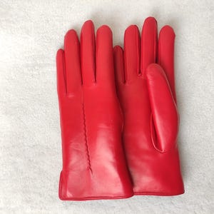 Op de afbeelding: Een paar felrode leren handschoenen. De handschoenen zijn glad en hebben een klassiek ontwerp, met een naad over de lengte van elke vinger. Ze liggen op een witte, getextureerde ondergrond.