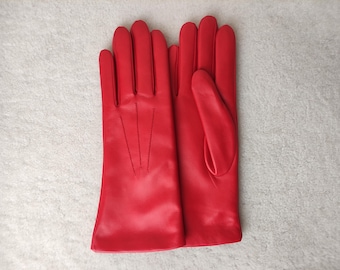 Puede incluir: Un par de guantes de color rojo vibrante con un acabado liso y tres líneas de costura verticales en el dorso de la mano. Los guantes tienen un corte clásico con una abertura recta en la muñeca y un pulgar separado, de diseño limpio.