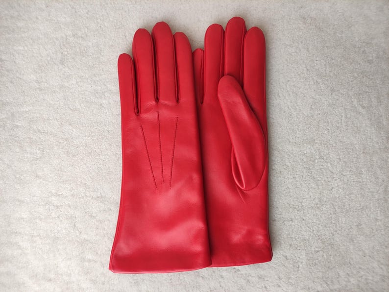 Peut inclure: Une paire de gants rouge vif avec une finition lisse et trois lignes de couture verticales sur le dos de la main. Les gants pr&eacute;sentent une coupe classique avec une ouverture droite au poignet et un pouce s&eacute;par&eacute;, au design &eacute;pur&eacute;.