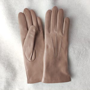 Peut inclure: Une paire de gants en cuir beige clair lisse. Un gant montre le c&ocirc;t&eacute; paume avec un pouce, tandis que l'autre affiche le dos avec trois lignes verticales cousues s'&eacute;tendant de la base des doigts vers le poignet.