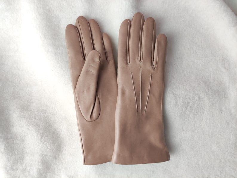 Peut inclure: Une paire de gants en cuir beige clair lisse. Un gant montre le c&ocirc;t&eacute; paume avec un pouce, tandis que l'autre affiche le dos avec trois lignes verticales cousues s'&eacute;tendant de la base des doigts vers le poignet.