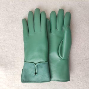 May include: A pair of green leather gloves. The gloves have a smooth texture and a contrasting suede cuff with a decorative keyhole detail. The gloves are designed for a comfortable fit and are likely for cold weather or fashion.
