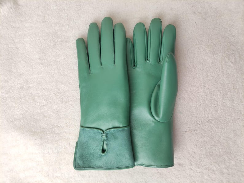 May include: A pair of green leather gloves. The gloves have a smooth texture and a contrasting suede cuff with a decorative keyhole detail. The gloves are designed for a comfortable fit and are likely for cold weather or fashion.