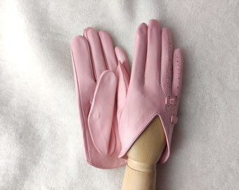 May include: A pair of light pink leather gloves featuring a smooth finish, a V-shaped wrist opening, and a row of small ventilation perforations along the side of the fingers. The gloves are displayed on a light-colored wooden hand form.