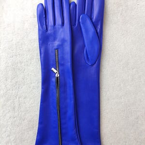 May include: A pair of long, vibrant blue leather gloves featuring a vertical silver-toned metal zip detail on the forearm of one glove. The gloves have a smooth finish and extend past the wrist, showcasing a sleek, fitted design.