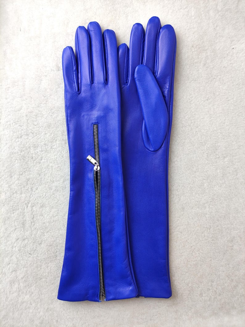 May include: A pair of long, vibrant blue leather gloves featuring a vertical silver-toned metal zip detail on the forearm of one glove. The gloves have a smooth finish and extend past the wrist, showcasing a sleek, fitted design.