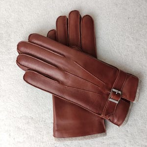 May include: A pair of brown leather gloves featuring three decorative stitched lines on the back of the hand, a wrist strap with a silver-toned metal buckle, and a light gray knit trim at the cuff edge, shown layered on top of each other.