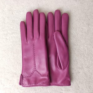 Puede incluir: Un par de guantes de cuero magenta. Los guantes tienen un dise&ntilde;o cl&aacute;sico con detalles de costura en la parte posterior de la mano. Los guantes se muestran sobre una superficie blanca y texturizada.