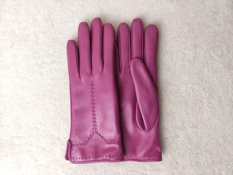 Puede incluir: Un par de guantes de cuero magenta. Los guantes tienen un dise&ntilde;o cl&aacute;sico con detalles de costura en la parte posterior de la mano. Los guantes se muestran sobre una superficie blanca y texturizada.