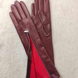 May include: A pair of deep burgundy leather gloves featuring a silver-toned metal zip along the side of the wrist. The interior lining of the zipped section is a vibrant red fabric, contrasting with the dark exterior leather finish.