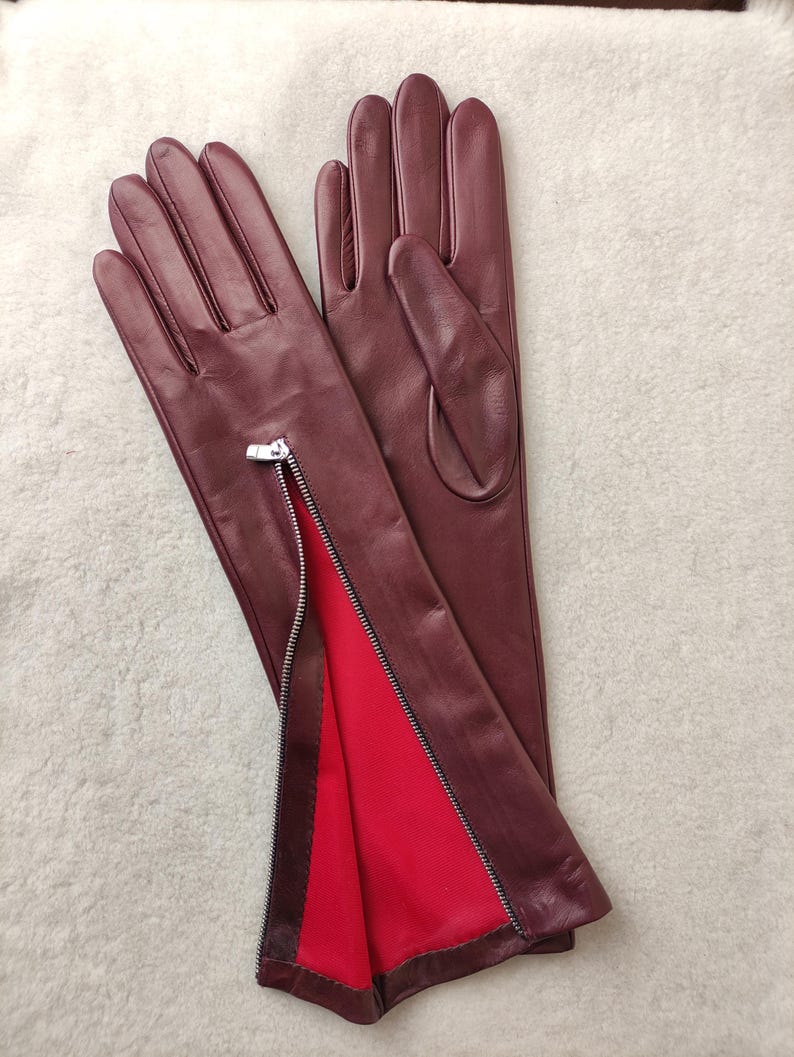 May include: A pair of deep burgundy leather gloves featuring a silver-toned metal zip along the side of the wrist. The interior lining of the zipped section is a vibrant red fabric, contrasting with the dark exterior leather finish.