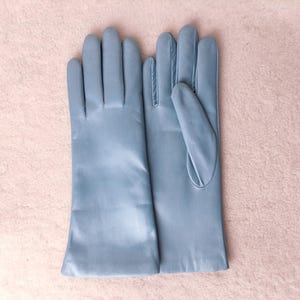 May include: A pair of light blue leather gloves with a smooth finish. The gloves feature a classic design with four fingers and a thumb, extending past the wrist. The material appears soft and uniform in color, showing clean stitching.