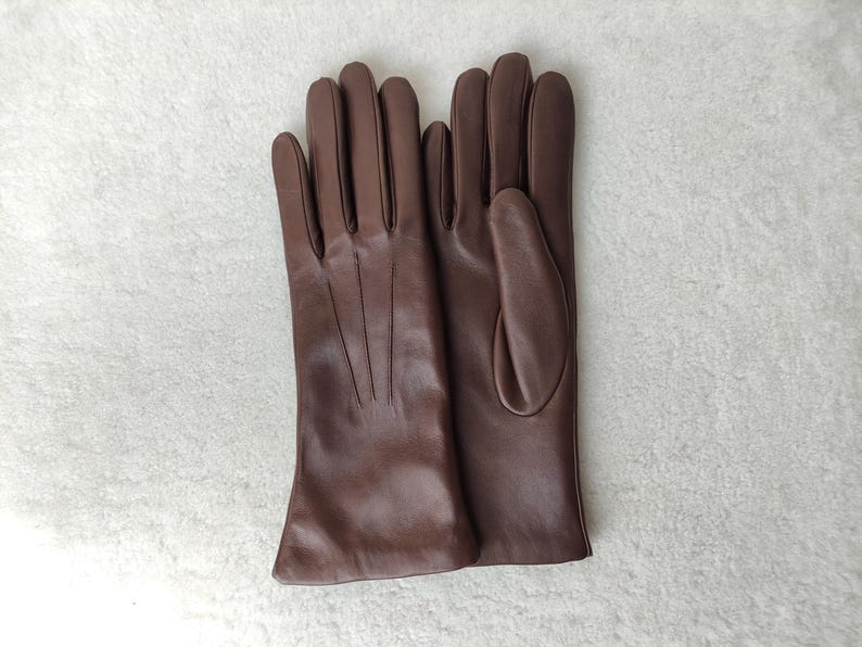 Peut inclure: Une paire de gants en cuir marron lisse pr&eacute;sentant trois lignes verticales cousues sur le dos de la main. Les gants ont une coupe classique avec une ouverture droite au poignet et un pouce positionn&eacute; sur le c&ocirc;t&eacute; du gant droit.
