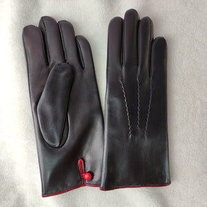 May include: A pair of dark brown leather gloves featuring a smooth finish, three decorative stitched lines on the back of the hand, and a small red button detail at the wrist with a contrasting red interior lining visible at the cuff.