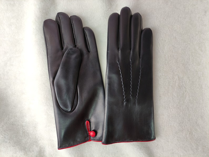 May include: A pair of dark brown leather gloves featuring a smooth finish, three decorative stitched lines on the back of the hand, and a small red button detail at the wrist with a contrasting red interior lining visible at the cuff.