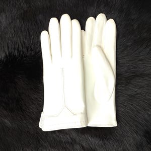 Puede incluir: Un par de guantes blancos impecables, probablemente de cuero, sobre una superficie oscura y texturizada. Los guantes son lisos, con costuras visibles y un dise&ntilde;o cl&aacute;sico. El fondo es de un negro profundo, proporcionando un fuerte contraste.