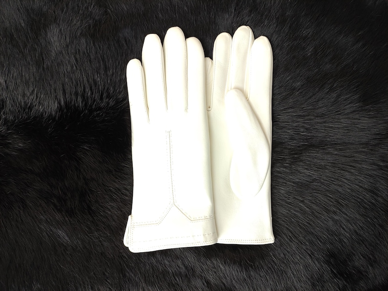 Puede incluir: Un par de guantes blancos impecables, probablemente de cuero, sobre una superficie oscura y texturizada. Los guantes son lisos, con costuras visibles y un dise&ntilde;o cl&aacute;sico. El fondo es de un negro profundo, proporcionando un fuerte contraste.